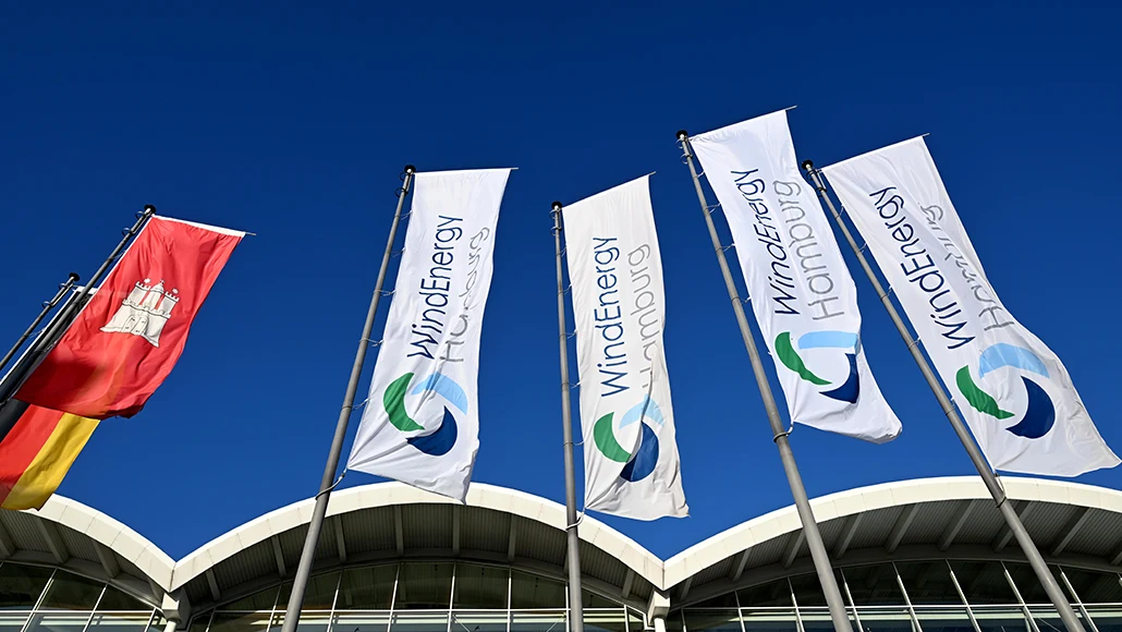 White WindEnergy Hamburg flags fluttering beside a modern curved-roof building under a clear blue sky.