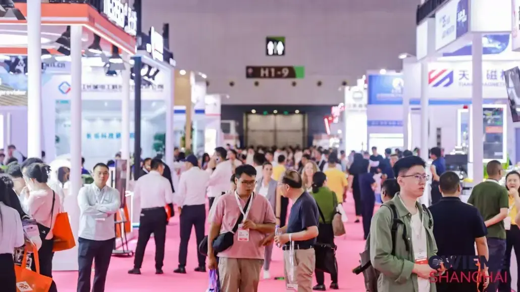 Busy trade show hall with attendees walking along a pink carpeted aisle and exhibition booths on both sides.