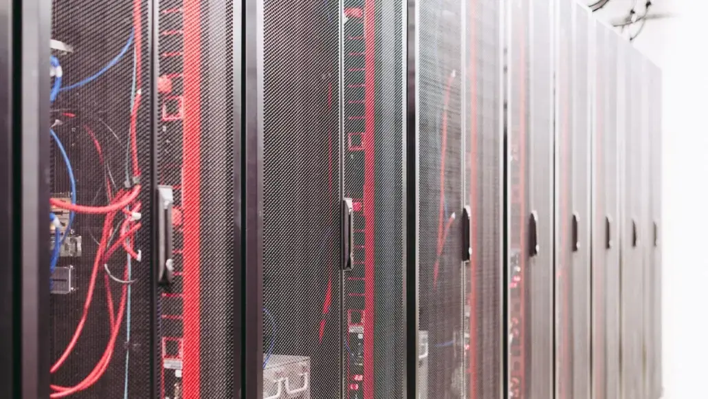 Row of tall black server racks with red and blue cables in a data center, behind mesh doors